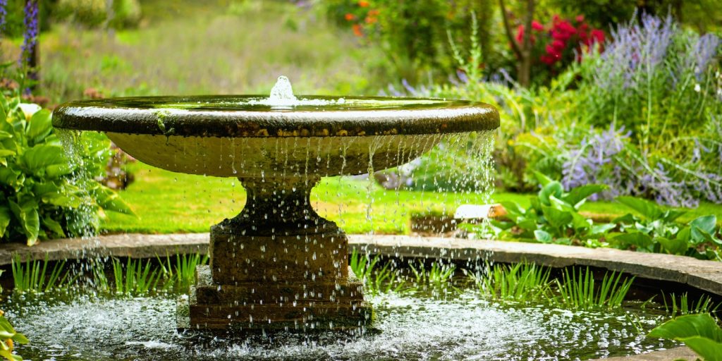 A water fountain in a garden, surrounded by grass and flowers